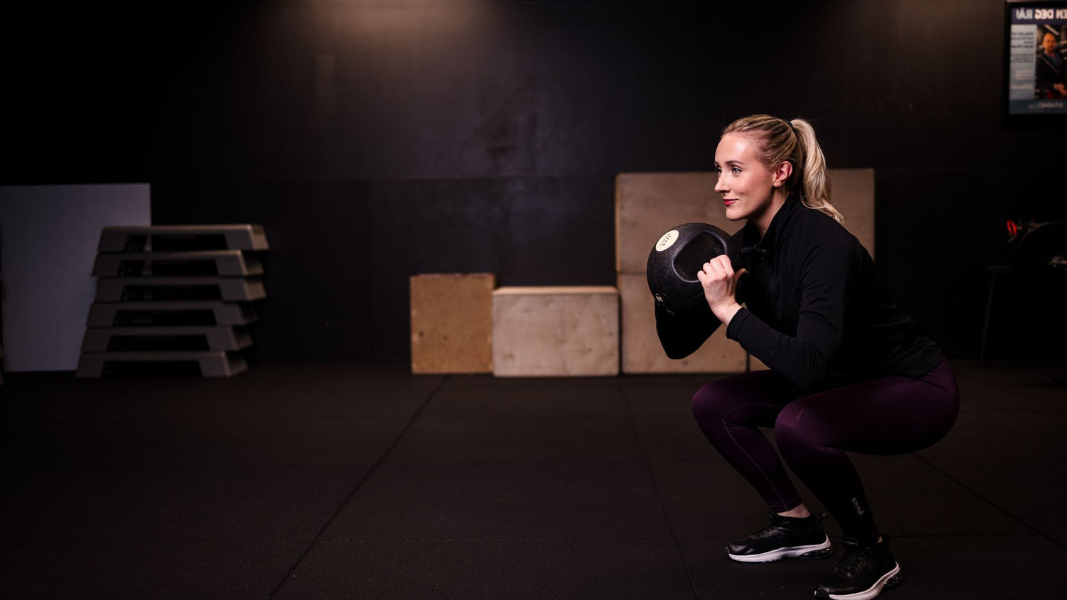 Elena Mellingen utfører en goblet squat med kettlebell på et treningssenter med dempet belysning.